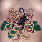 A speckled black scorpion, sitting inside a yellow and pink chrysanthemum in a japanese style. It is tattooed on a woman's sternum by Harriet Street at Cult Classic Tattoo in Romford, Essex